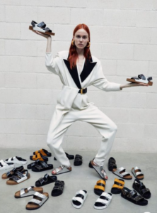 Red-headed woman holding two sandals next to a pile of sandals
